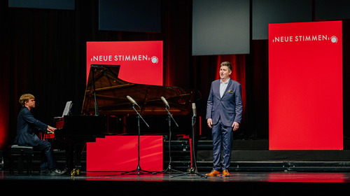 Kieran Carrel, Teilnehmer des NEUE-STIMMEN-Wettbewerbs 2022, steht beim Vorsingen in der Endrunde in Gütersloh auf der Bühne und singt, neben ihm ist ein Pianist am Flügel zu sehen.